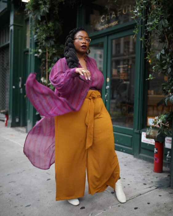 Looks coloridos aesthetic; mujer afroamericana, morena, con cabello negro peinado en una trenza gruesa, lentes de aumento retro, labial rojo vino, blusa de gasa rosa con rayas verticales, pantalón ligero amarillo mostaza, botines blancos