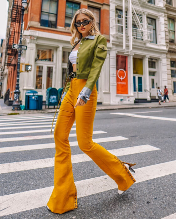 Looks coloridos aesthetic; mujer caminando en la calle en paso peatonal, cabello rubio, mediano, ondulado, suelto y despeinado, lentes de sol rectangulares, crop top blanco, chamarra verde militar, pantalones de mezclilla acampanados amarillo anaranjado, botines blancos