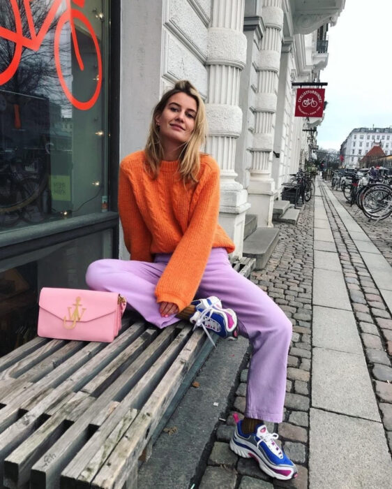 Looks coloridos aesthetic; mujer sonriendo sentada en banca de madera en la calle, cabello rubio, mediano, lacio, suelto y despeinado, atuendo de colores, suéter tejido anaranjado, pants morado lila, tenis chunky de duela gruesa azul fuerte y rosa mexicano, bolsa de mano en tonos pastel con cadena dorada