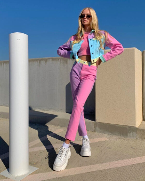 Looks coloridos aesthetic; mujer rubia parada en estacionamiento con atuendo de colores, cabello rubio, lacio, largo y suelto, crop top rosa, jeans, pantalones de mezclilla  en tonos pastel, lila, amarillo y azul, chamarra a juego, tenis chunky de suela gruesa y plataforma de color blanco, lentes de sol cuadrados