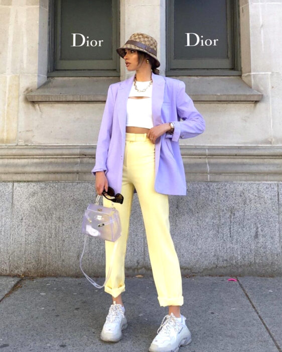 Looks coloridos aesthetic; mujer de cabello castaño peinado, con gorro de pescador café a cuadros, crop top de vestir blanco, pantalón amarillo pastel, blazer, saco morado lila, bolsa de mano transparente, lentes de sol cuadrados, tenis chunky grandes color blanco, frente a tienda Dior