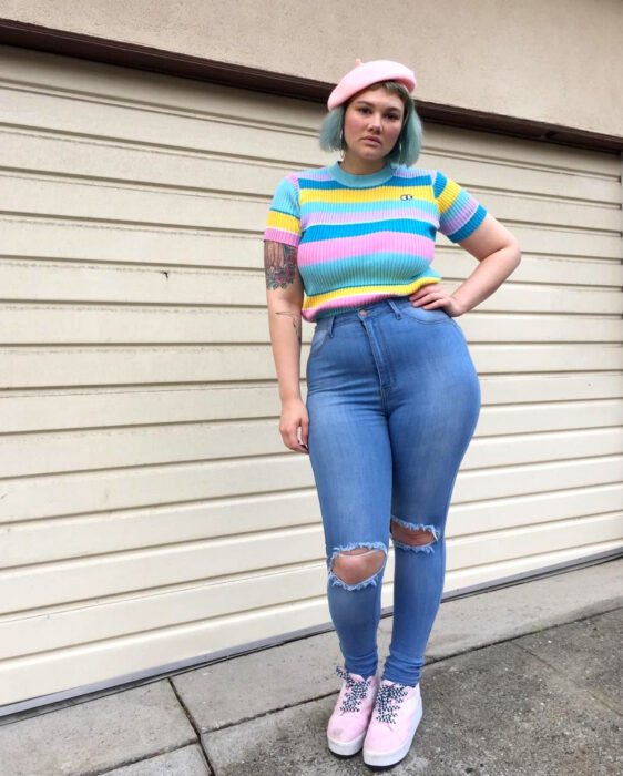 Looks coloridos aesthetic; mujer plus size con la mano en la cintura, atuendo de colores, boina francesa rosa pastel, blusa de manga corta azul fuerte y clarito, amarillo, jeans, pantalones de mezclilla desgastados, rotos de las rodillas, tenis de tonos pastel, cabello corto de hinguito con tinte fantasía, tatuaje de rosas en el brazo