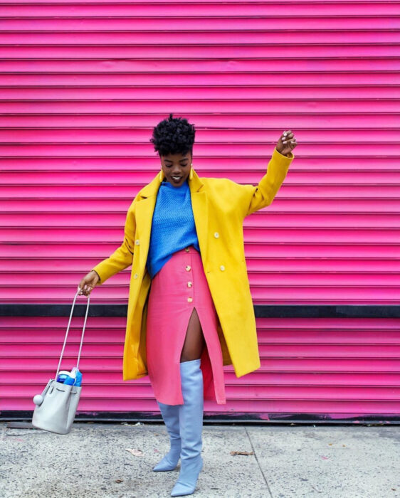 Looks coloridos aesthetic; mujer afroamericana de piel morena, cabello chino, sonriendo, vestida con atuendo de colores, blusa azul, falda lápiz rosa con abertura en la pierna, botas largas a la rodilla grises, blazer abrigo amarillo, bolsa de mano y aretes largos