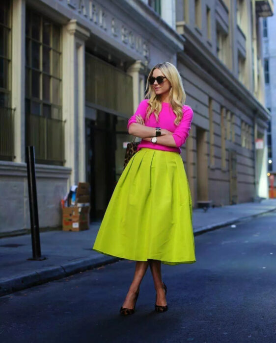 Looks coloridos aesthetic; chica posando en la calle, cabello rubio, largo, peinado suelto y ondulado, lentes de sol oscuros, grandes y rectangulares, suéter con manga 3/4 color rosa mexicano, falda larga corte A verde neón, zapatillas animal print, reloj de mujer, pulsera, bolsa de mano