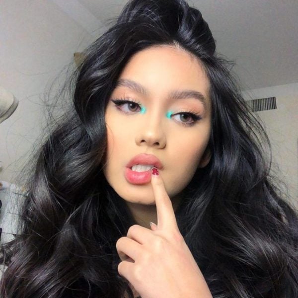 Chica asiática con cabello largo y suelto se lleva el dedo a los labios mientras usa un maquillaje suave de ojos con el lagrimal en color azul turquesa 