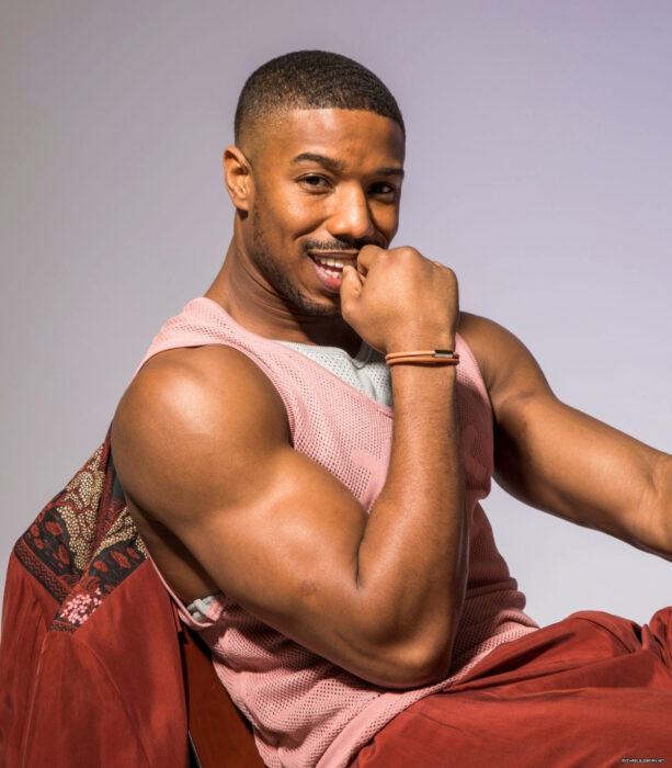 Actor Michael B. Jordan sentado, con una playera blanca y rosa de tela de red, pantalón y saco rojo, mostrando sus brazos musculosos, sonriendo con mano en la boca