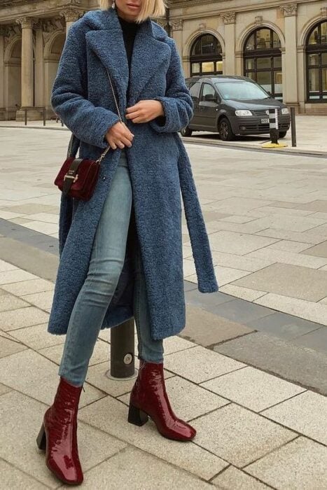 Chica usando jeans, blusa negra, abrigo de peluchito color azul largo, con un bolso y botines color vino