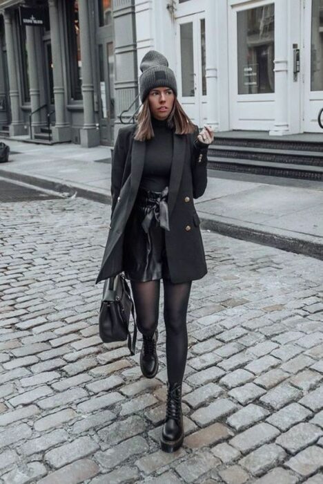 Chica usando botines, con medias, mini falda de cuero, suéter de cuello alto y abrigo largo con bolsa de mano todo en color negro, con gorrito color gris rata