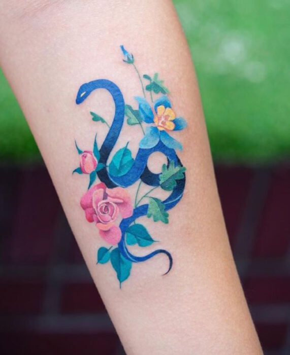 Tatuaje de serpiente en tinta de color azul, y flores sobre el área del antebrazo