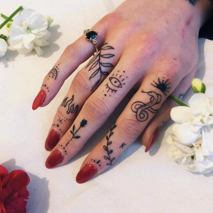 Manos bonitas de mujer con manicura, uñas largas en forma de almendra con esmalte color rojo, dedos con tatuajes de ojos, llamas, flores, serpiente, hojas y sol, anillo de piedra negra, flor blanca y roja