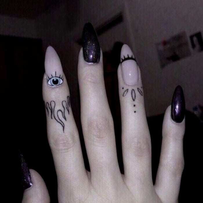 Hübsche Frauenhände mit Maniküre, lange Stiletto-Nägel mit blassrosa und schwarzer Glitzerpolitur, Augen- und Wimpernmuster, Finger mit Stammes-Tattoos und Flammen