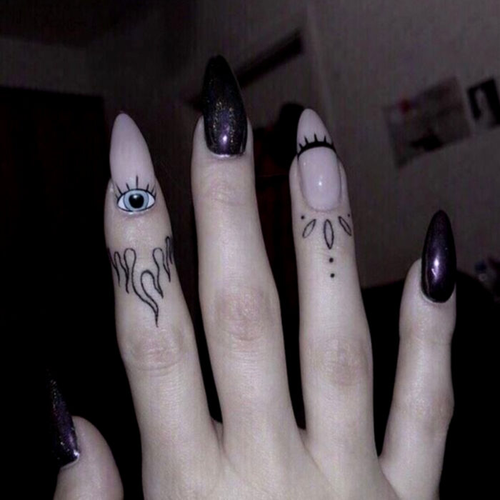 Manos bonitas de mujer con manicura, uñas largas stiletto con esmalte de color rosa pálido y negro con brillos, diseño de ojos y pestañas, dedos con tatuajes tribales y flamas