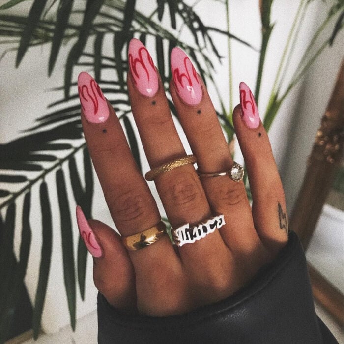 Manos bonitas de mujer con manicura, uñas largas en forma de almendra con esmalte color rosa con diseño de flamas rojas, dedos con tatuajes de puntos y rayos, y anillos dorados