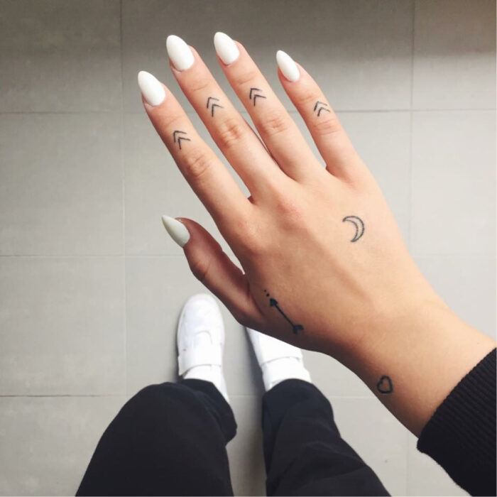 Manos bonitas de mujer con manicura, uñas lardas en forma de almendra con esmalte color blanco, dedos con tatuajes de flechas minimalistas, tattoo de luna y corazón en la mano y en la muñeca, tenis blancos