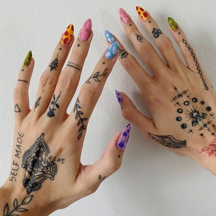 Manos bonitas de mujer con manicura, uñas largas stiletto con esmalte de colores verde, amarillo, rosa, azul y morado con lunares, manos y dedos con tatuajes de puntos, flechas, rosas, triángulos, enredaderas, alas, fases de la luna, montañas, ojos y frases