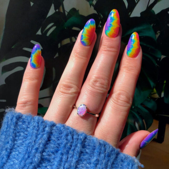 Diseños bonitos de manicura coloridos; uñas largas en forma de almendra pintadas con esmaltes de colores jipis del arcoíris, morado, azul fuerte y claro, verde, amarillo, anaranjado y rojo, anillo de piedra rosa