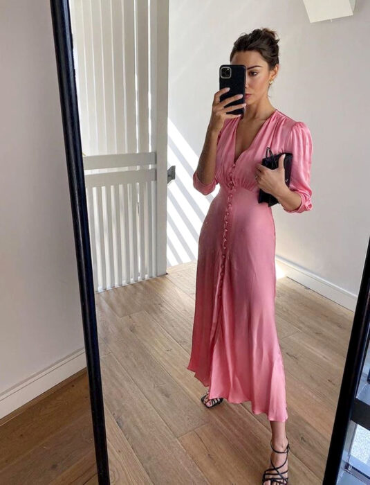 Mujer tomándose una selfie con su celular frente al espejo, vestido rosa largo y liso, de mangas a los codos, escote en v y botones al frente, peonado de chongo recogido y cabello castaño, bolsa de mano negra, sandalias de tacón negras