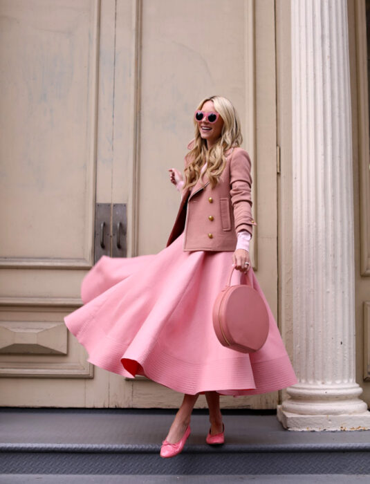 Mujer sonriendo en la calle frente a una puerta grande, cabello rubio, largo, ondulado y suelto, lentes de sol de Barbie, vestido rosa largo y con vuelo, saco de vestir, flats, bolsa redonda de mano