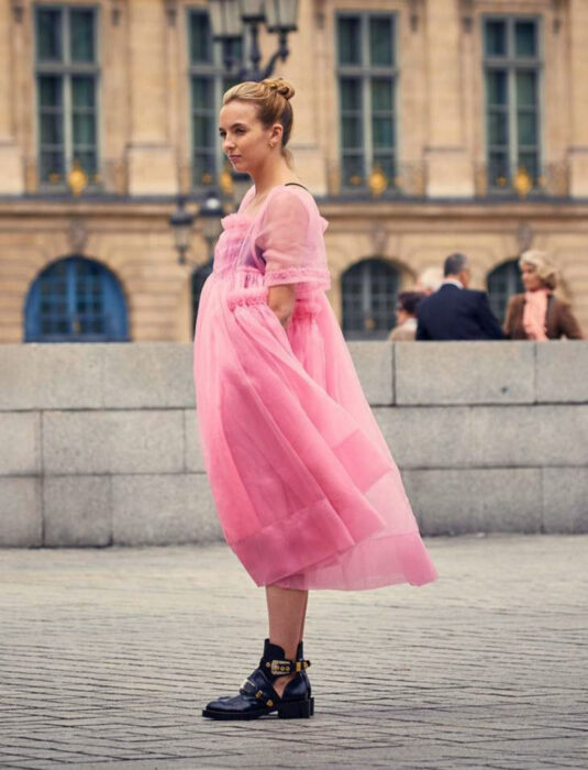 Mujer en una plaza, cabello rubio con peinado de chongo alto, vestido de tela de tul transparente color rosa, botines negros con estoperoles