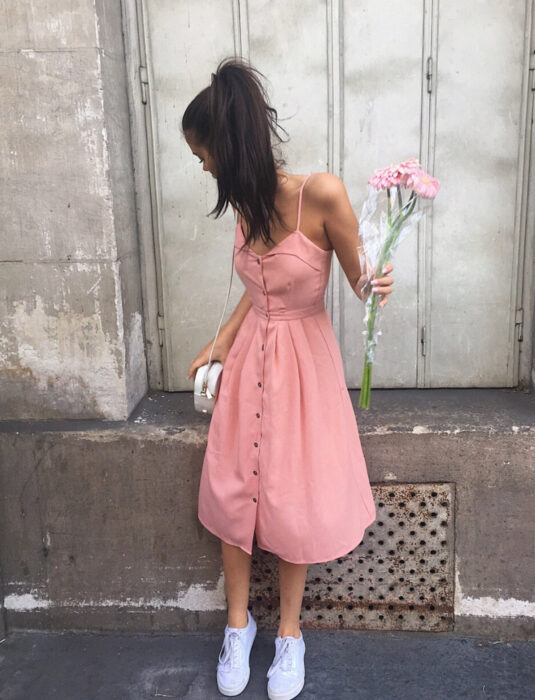 Mujer morena con vestido rosa de tirantes, botones y debajo de las rodillas, con un ramo de flores, bolsa color marfil, tenis blancos, cabello castalo, largo, lacio peinado con una cola de caballo alta