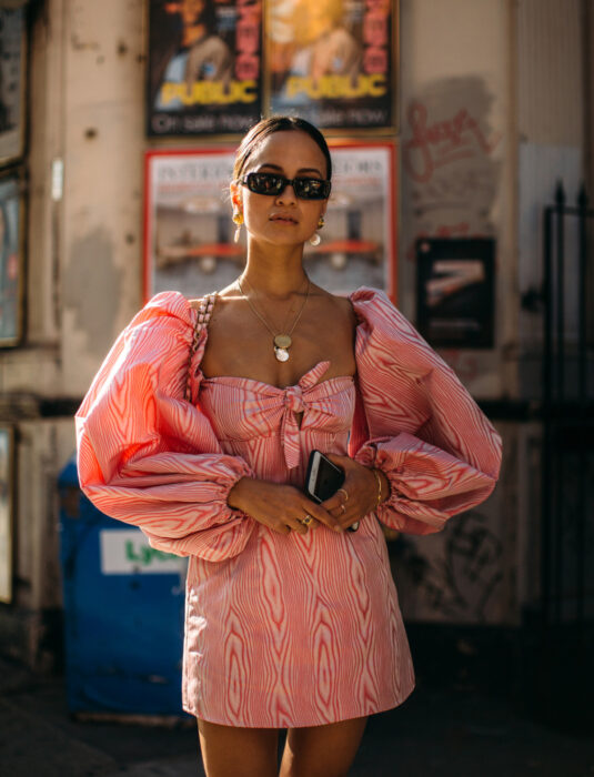 Mujer en la calle, cabello castaño oscuro, peinada con una cola baja, lentes de sol oscuros rectangulares, aretes y collares dorados, vestido color rosa con blanco, mangas abombadas, escote cuadrado, con el celular en la mano