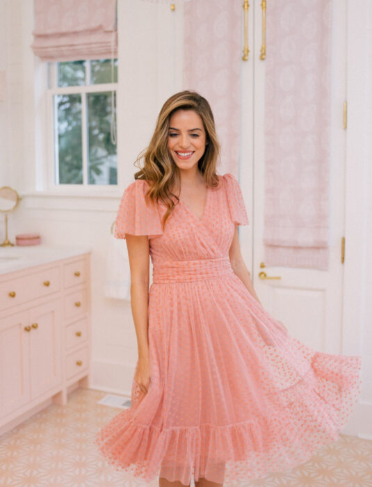 Mujer sonriendo, cabello rubio, mediano, ondulado y suelto, con vestido femenino rosa de tela de tul con lunares