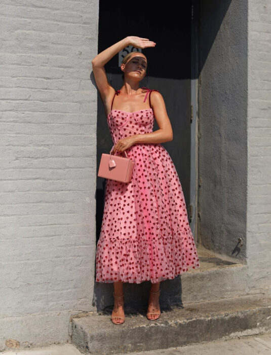 Mujer en la calle tapándose el rostro del sol, con vestido rosa de tirantes, con estampado de corazones, corset, tela de tul, largo hasta los tobillos, sandalias de tacón nude, bolsa de mano rectangular