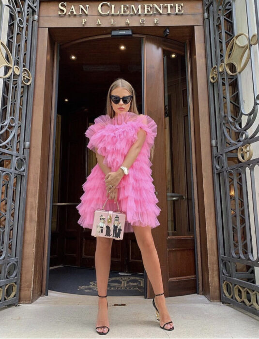 Chica frente a puerta de madera, cabello castaño rubio, lacio y largo, con lentes de sol oscuros en forma de corazón, vestido rosa de holanes, bolsa de mano rectangular, sandalias negras de tacón dorado, reloj de mujer