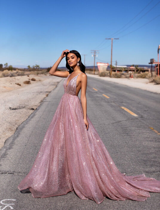 Mujer morena en medio de la autopista, de cabello castaño oscuro, lacio y debajo de los hombros, vestido de fiesta rosa brillante, largo, de tirantes, escote en v, aretes grandes