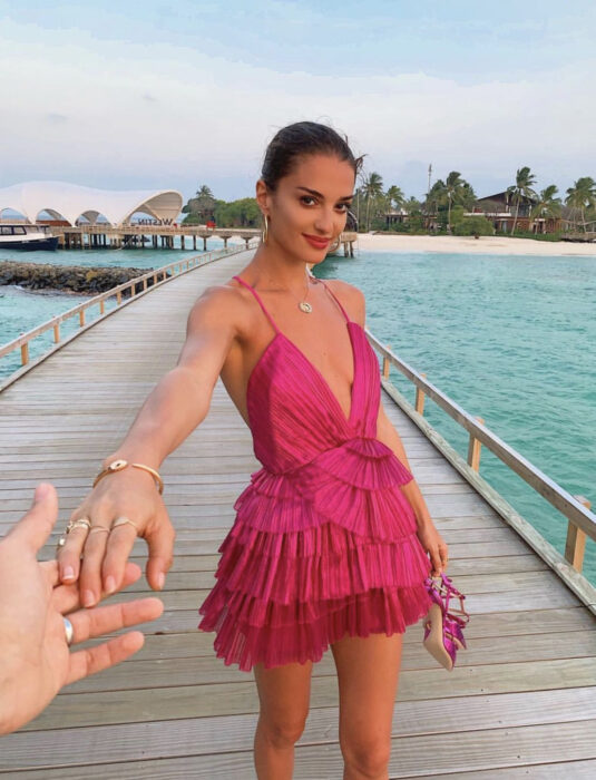 Mujer morena en la playa, en puerto sobre el mar, cabello castaño oscuro, peinado en un chongo bajo, vestido playero rosa mexicano con holanes, tirantes, escote en v, sandalias en las manos