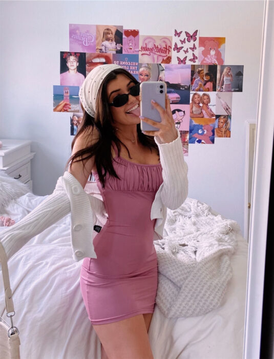 Mujer tomándose una selfie frente al espejo con el celular, minivestido rosa de tirantes, ceñido al cuerpo, paliacate en el cabello castaño, lacio y suelto, suéter tejido blanco, lentes de sol ovalados negros, pared llena de posters de artistas y películas