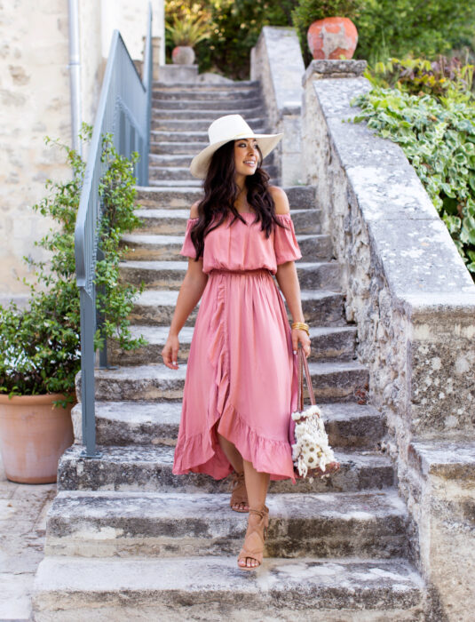 Mujer bajando escaleras de piedras con plantas alrededor, cabello castaño oscuro, ondulado, largo y suelto, con vestido rosa playero sin hombros y hasta los chamorros, sandalias de piso café, bolsa de mano, sombrero blanco