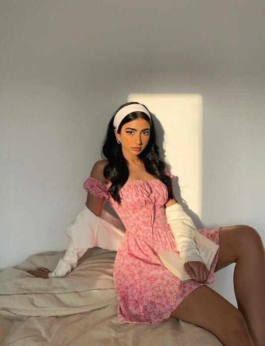Mujer morena sentada en la cama leyendo un libro, de cabello castaño oscuro, ondulado, largo y suelto, con una bandana blanca, minivestido campesino rosa de flores, sin hombros, suéter color beige
