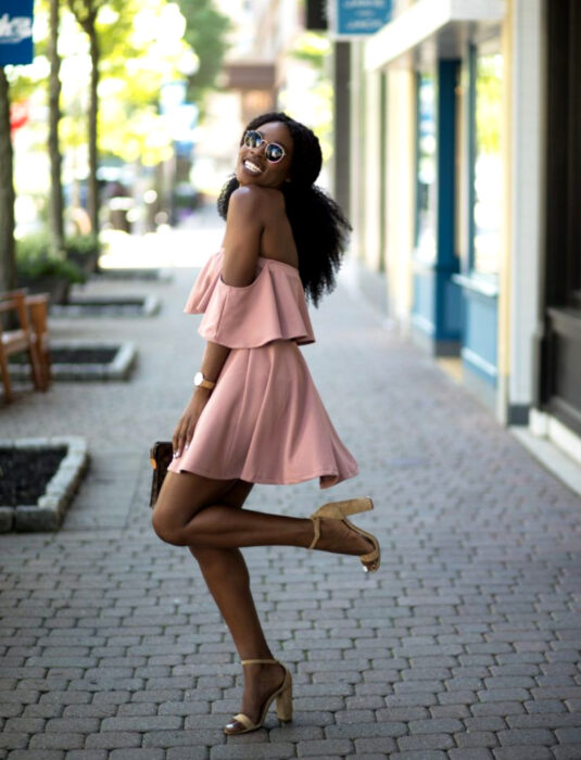 Chica morena sonriendo en la calle, cabello chino, negro, afro, esponjado peinado con media cola, lentes de sol oscuros, minivestido rosa sin hombros, tacones color camel, reloj de mujer