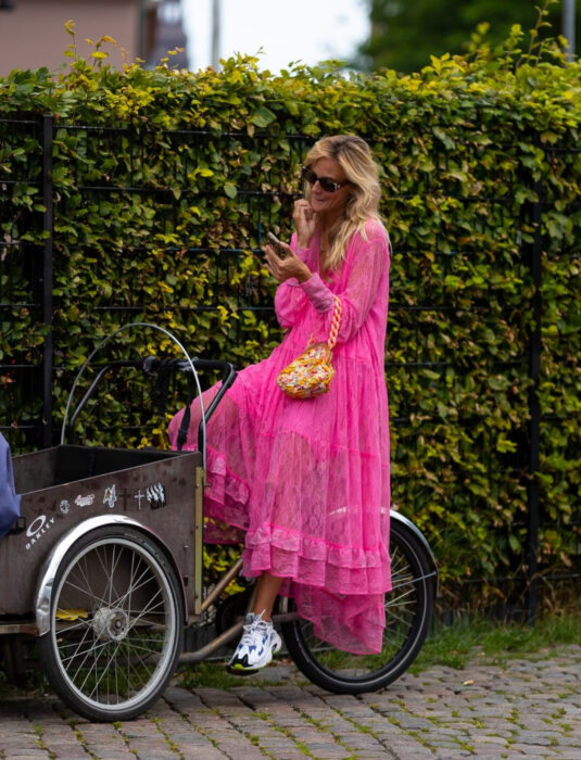 Mujer en triciclo frente a un arbusto en la calle, mirando su celular, cabello rubio, ondulado, suelto, despeinado, mediano, con vestido rosa neón de encaje y holanes hasta los tobillos, tenis skechers blancos con detalles azules, negros y berdes, bolsa de mano amarilla con cadena gruesa, lentes de sol rectangulares