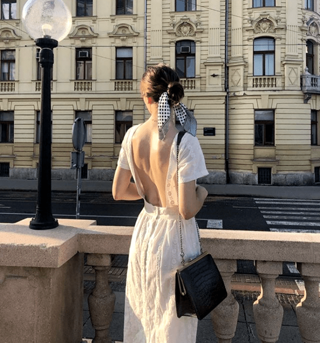 chica de cabello oscuro usando un vestido blanco de manga corta con escote en la espalda, bolso de  mano negro con correa de cadena