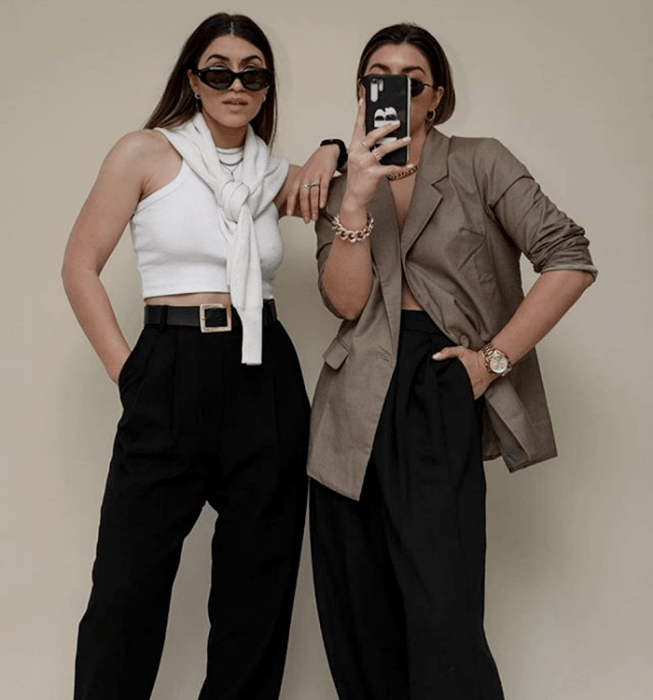 dos chicas de cabello castaño con lentes de sol, top blanco sin mangas de cuello alto, blazer café oversized, pantalones negros de vestir y suéter blanco