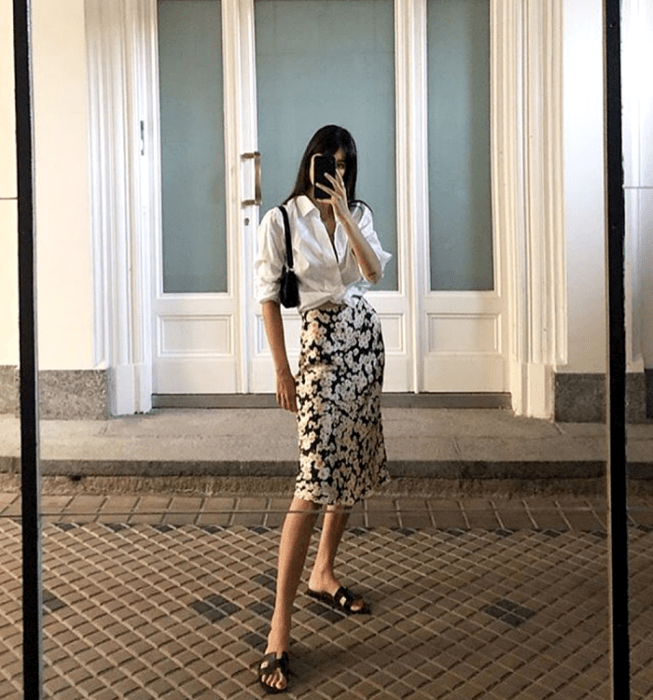 chica de cabello castaño usando una camisa de vestir blanca, falda de satén negra con flores blancas, sandalias negras de piso y bolso de mano negro