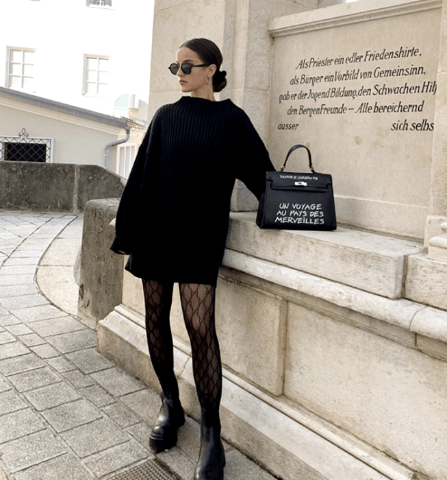 chica de cabello negro con lentes de sol, suéter oversized negro, minifalda negra, medias negras de red y botas de plataforma negras, bolso de mano  negro con letras blancas