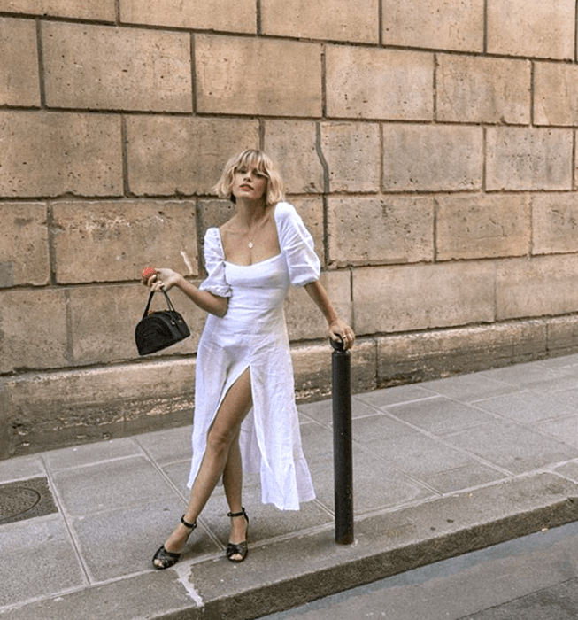 chica rubia usando un vestido blanco de mangas cortas abombadas, escote cuadrado y apertura en la pierna, bolso de mano negro mini