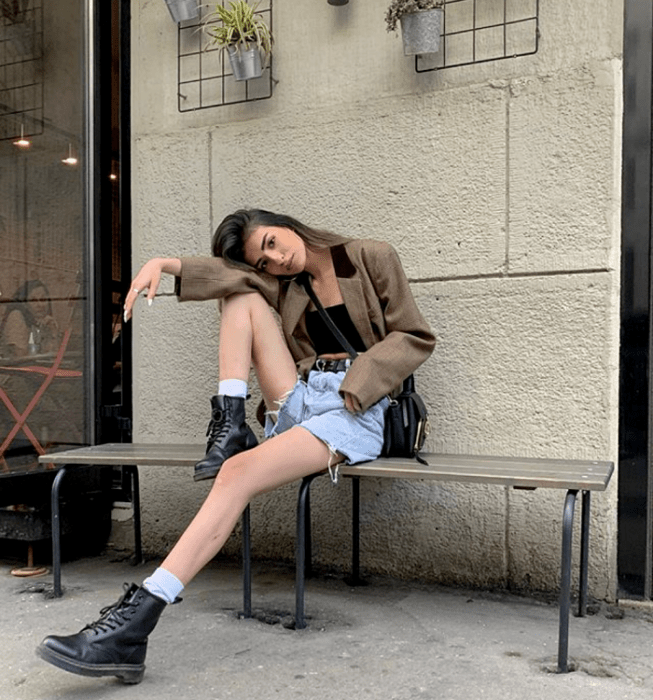 chica de cabello castaño usando un crop top negro sin mangas, blazer café con cuello negro, shorts de mezclilla, botines negros de piel y bolso negro crossbody bag