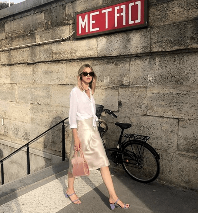 chica rubia usando lentes de sol, camisa de vestir blanca, falda de satén color champaña, sandalias de tacón color lila y bolso de mano rosa mini