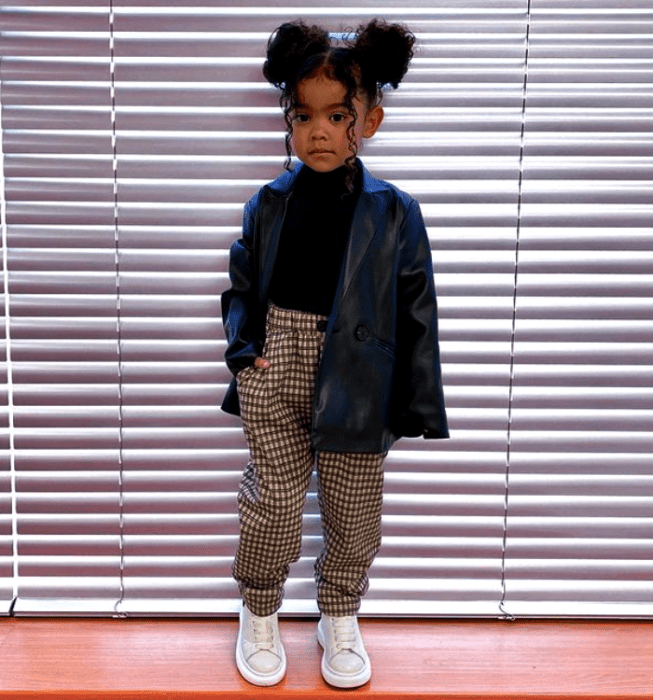 niña morena de cabello chino usando una blusa negra, chamarra de cuero negra, pantalones cafés, tenis blancos