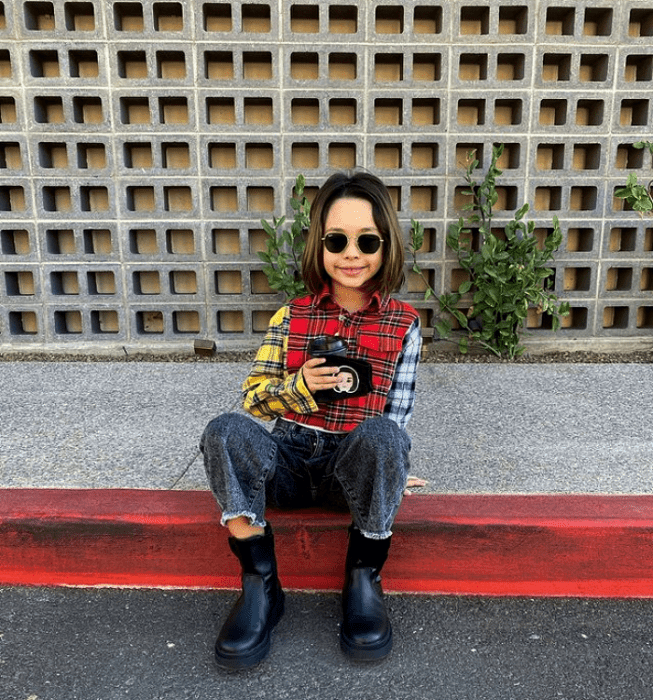 niña de cabello castaño usando lentes de sol, suéter de manga larga rojo, amarillo y azul, botines negros, pantalones grises