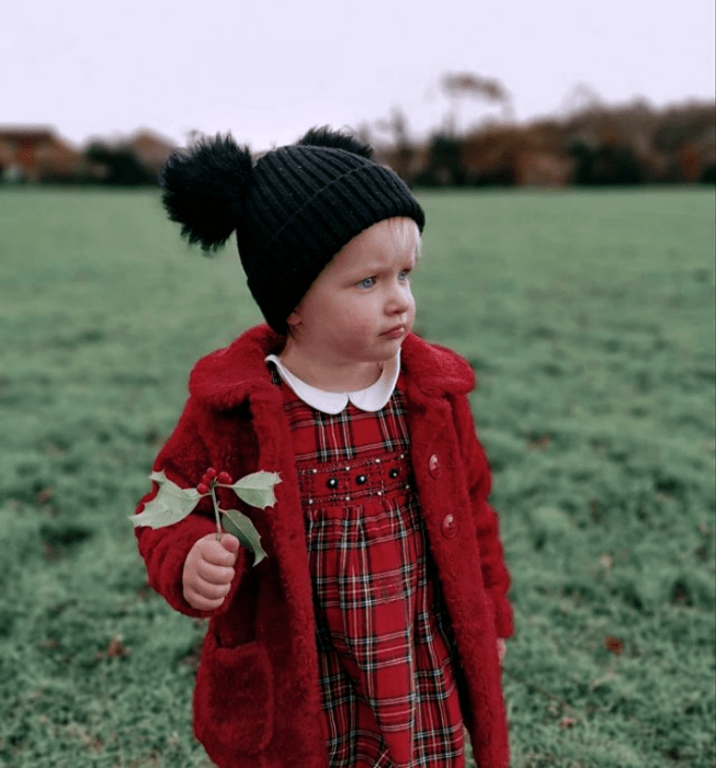 niña rubia de ojos azules usando un gorro negro tejido, vestido rojo de cuadros, abrigo largo rojo