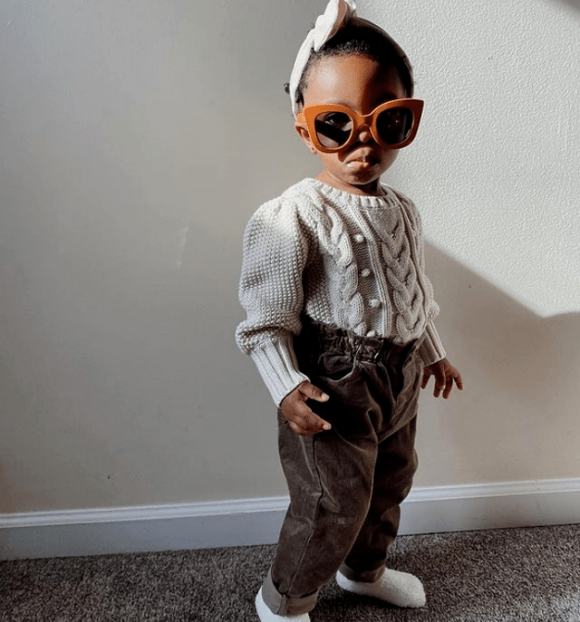 niña bebé morena con lentes de sol, suéter tejido blanco, pantalones cafés, calcetines blancos