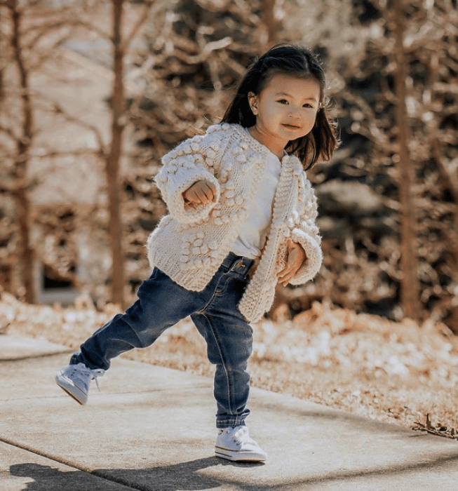 niña de cabello corto usando una camiseta blanca, suéter cárdigan beige, tenis blancos y pantalones de mezclilla