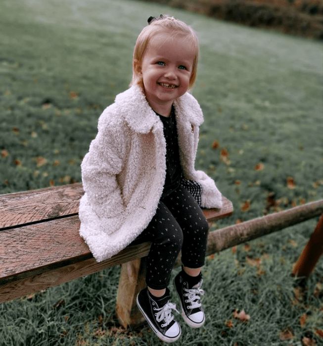 niña de cabello rubio con ojos azules, usando una sudadera negra con lunares blancos, pantalon negro con lunares blancos, tenis converse negros y abrigo teddy color beige