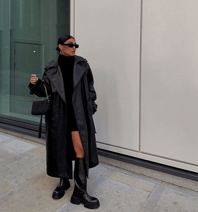 chica de cabello oscuro con lentes de sol, vestido mini negro de cuello alto, abrigo largo de cuero y botas largas de suela gruesa de cuero negro