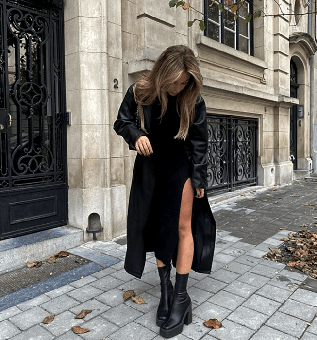 chica rubia usando un abrigo de cuero largo, vestido tejido negro con apertura en la pierna, botas de cuero de suela gruesa y bolso de mano negro